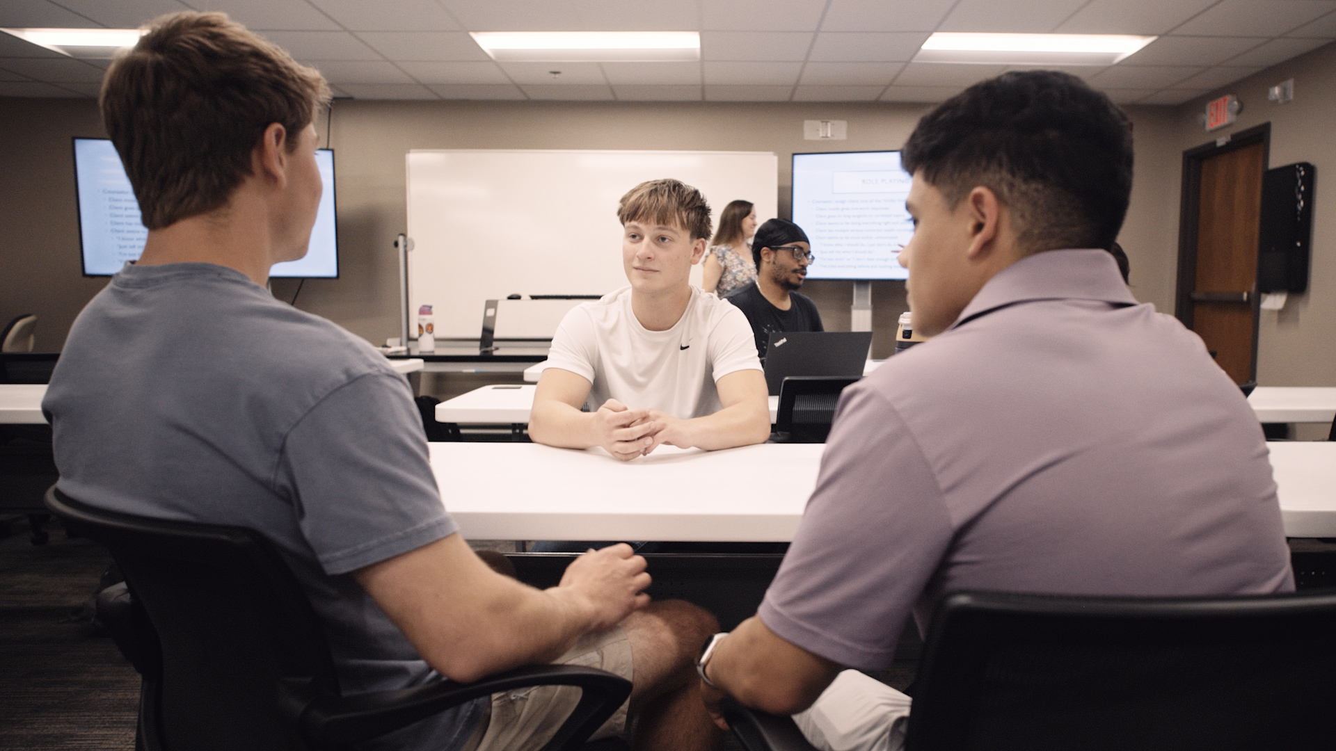 Three students sit at a table, with two on one side and one across. They are practicing health counseling.