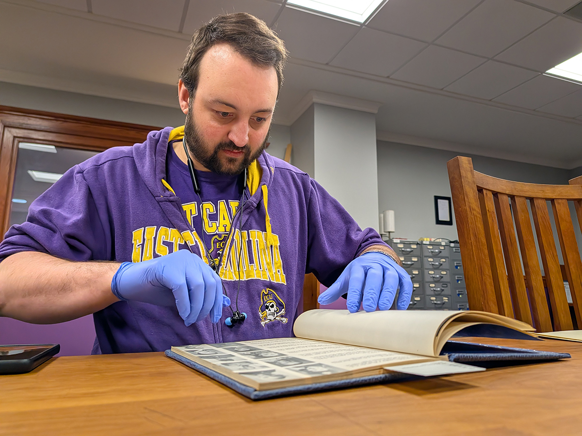 A college student in a hoodie and blue rubber gloves thumbs through a historic book at a table.