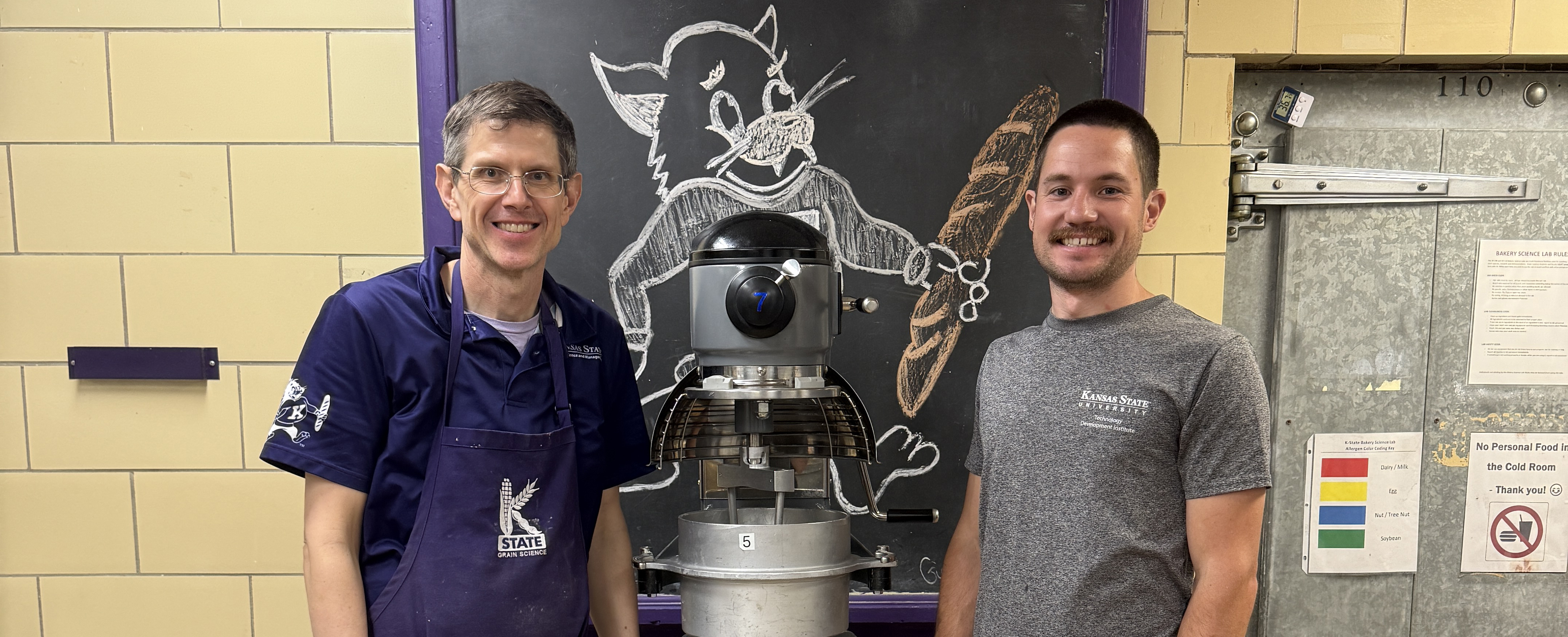 Two men stand on either side of an industrial mixer in front of a chalk drawing of Willie the Wildcat holding a baguette.