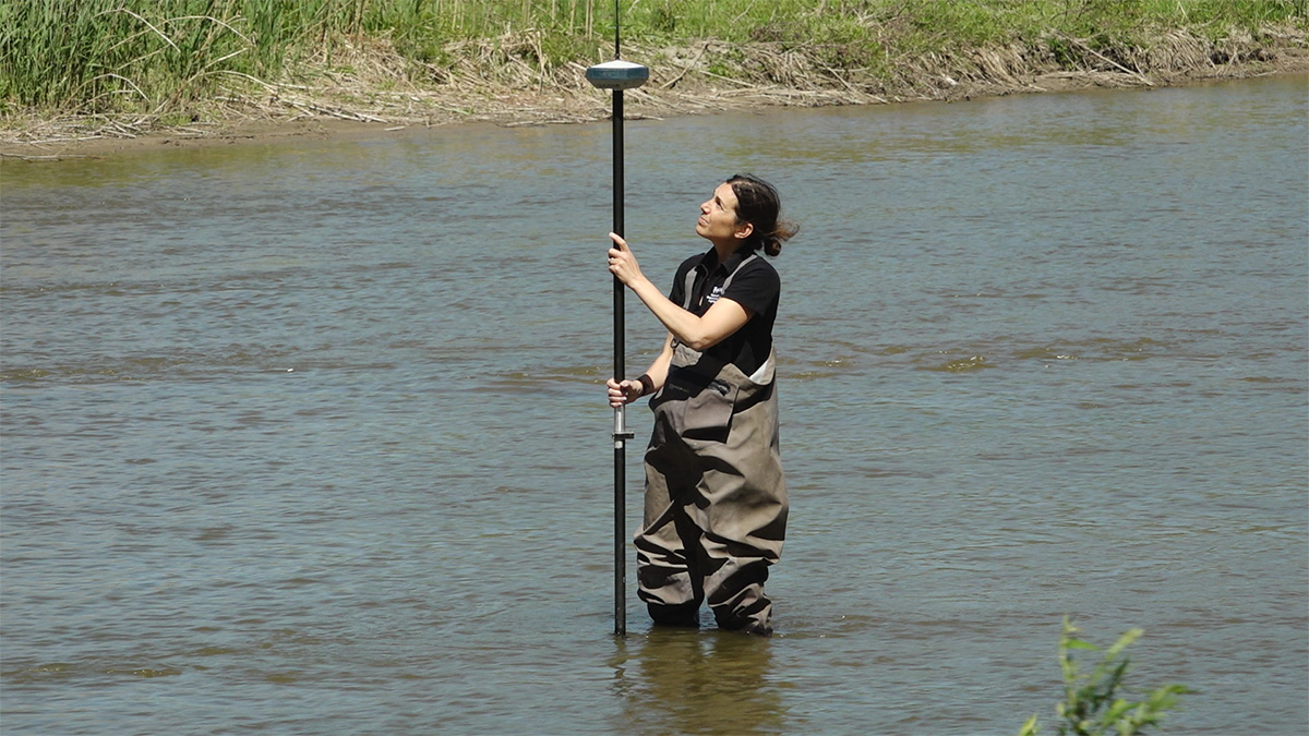 A woman in a black t-shirt and waders holds a long vertical measuring device in the middle of shin-length water.