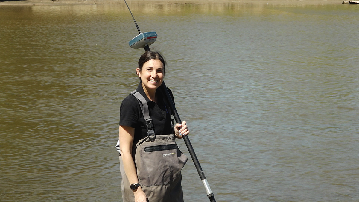 A woman wearing a black t-shirt and waders stands in shin-length water holding a vertical measuring device.