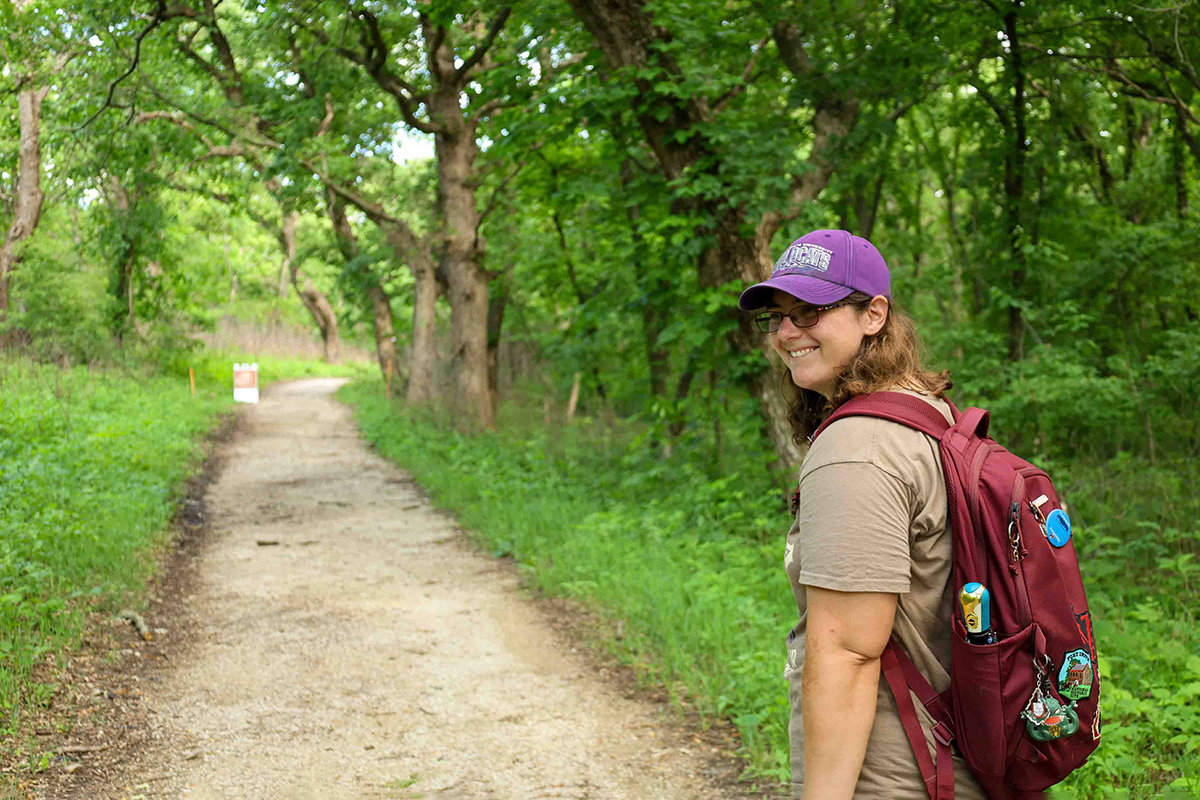 A doctoral student in a t-shirt, backpack and ballcap begins a hike in a wooded prairie trail.