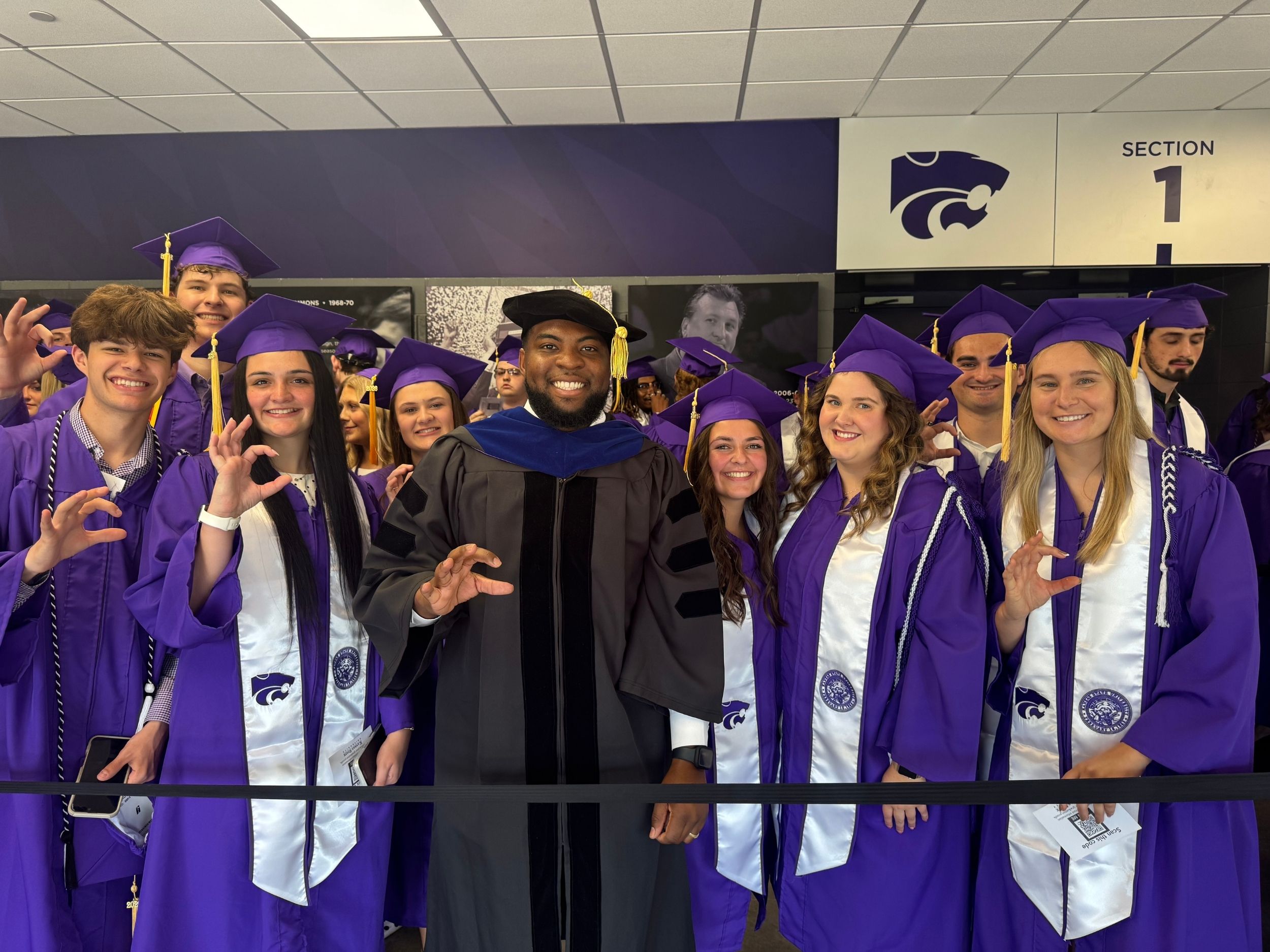 A professor in graduation regalia stands and smiles with a group of graduates in purple caps and gowns 