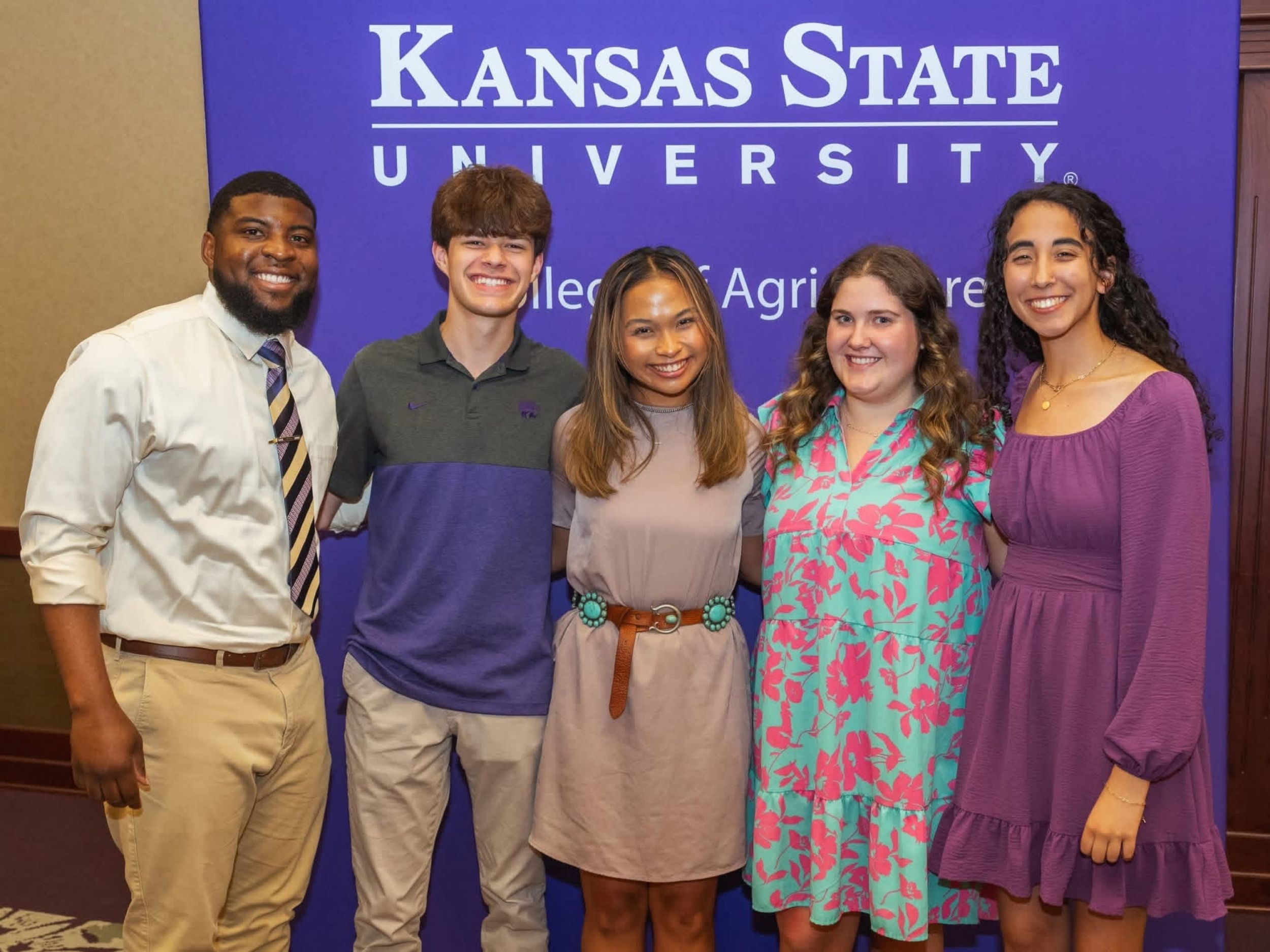 A professor in a white shirt and tie stands and smiles with four students in front of a purple Kansas State University background. 