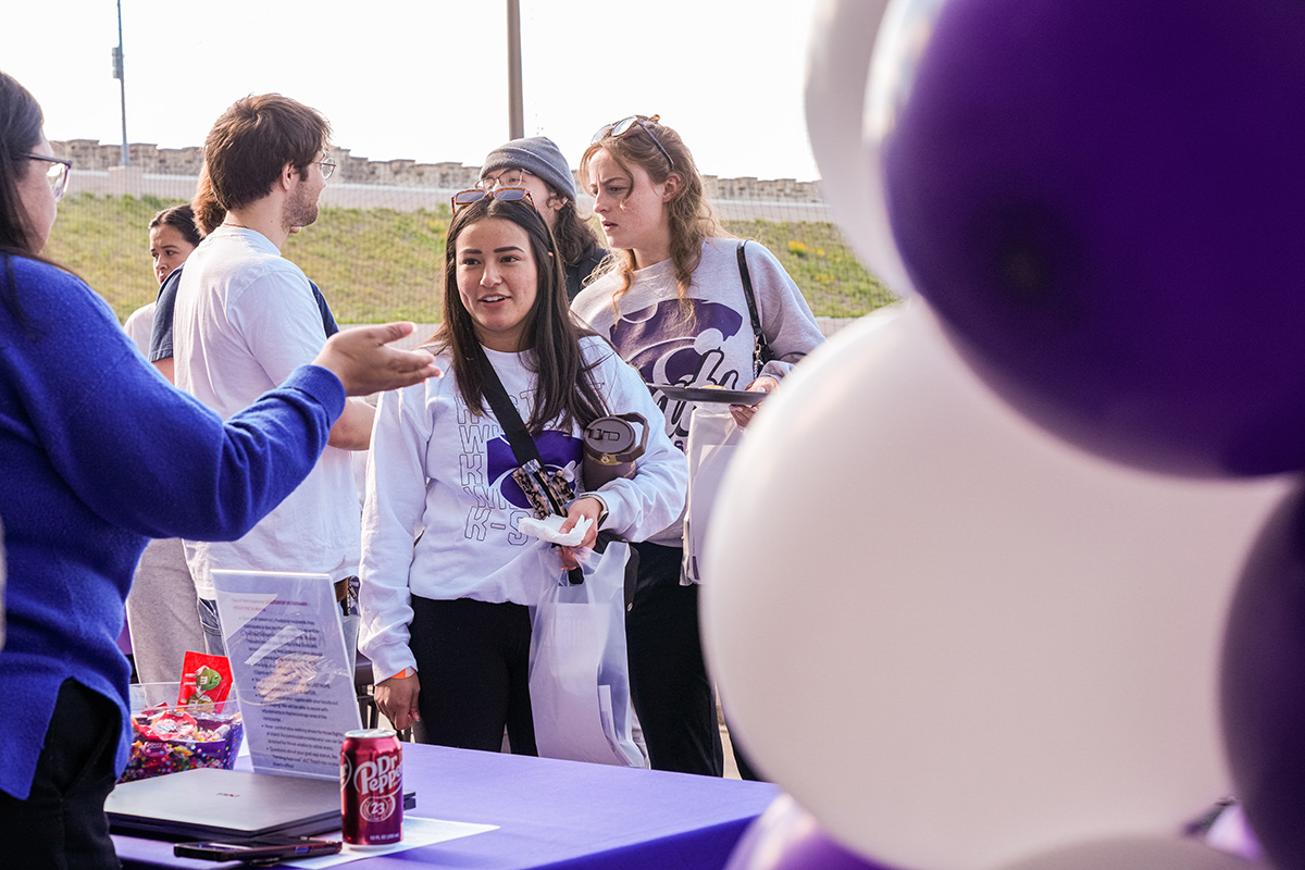 Student wearing white K-State hoodie and black crossbody bag smiles while receiving purple giveaway items from person in blue shirt at table covered with purple tablecloth. White balloons, red snacks, and water bottles sit on a table with grassy hills and concrete walls visible in the background under the overcast sky.