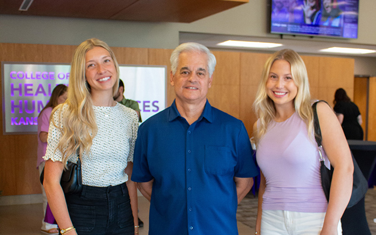 Three people pose together with woman on left wearing white patterned blouse and black pants, man in center wearing blue button-up shirt and beige pants, and woman on right wearing light pink sleeveless top and white pants.