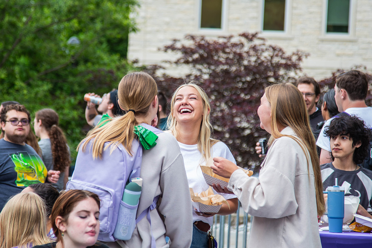 Three students stand talking and laughing outdoors with center woman in white shirt and gray cardigan holding plate of food, flanked by woman in light blue button-up with green scarf and woman in beige cardigan. A crowd of students visible in the background with limestone buildings, dark red foliage trees, and green trees behind them.