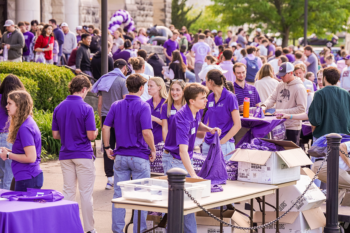 Large crowd gathers outdoors at events with volunteers in purple K-State t-shirts staffing tables covered with purple tablecloths displaying giveaway items and materials. Limestone building, green shrubs, purple and white balloons, and trees visible in background with attendees wearing a mix of purple and casual clothing.