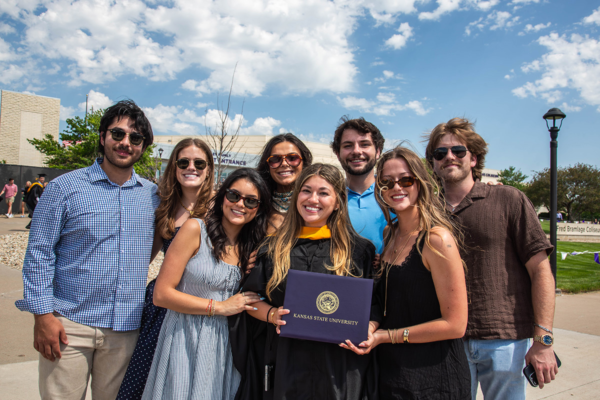 Group of eight people pose outdoors with graduates wearing black dresses and holding a purple diploma cover with gold seal, surrounded by family and friends wearing sunglasses and casual clothing.