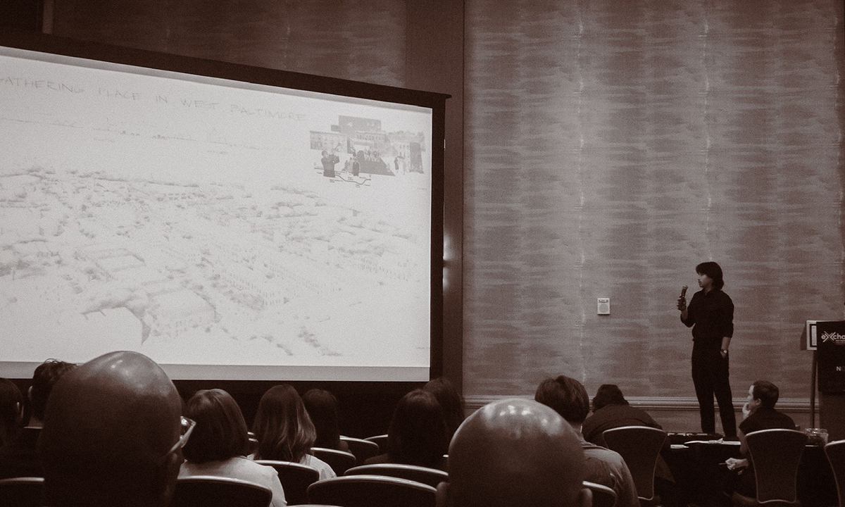 An architecture graduate student presents his portfolio on a stage in front of a large display in a conference room.