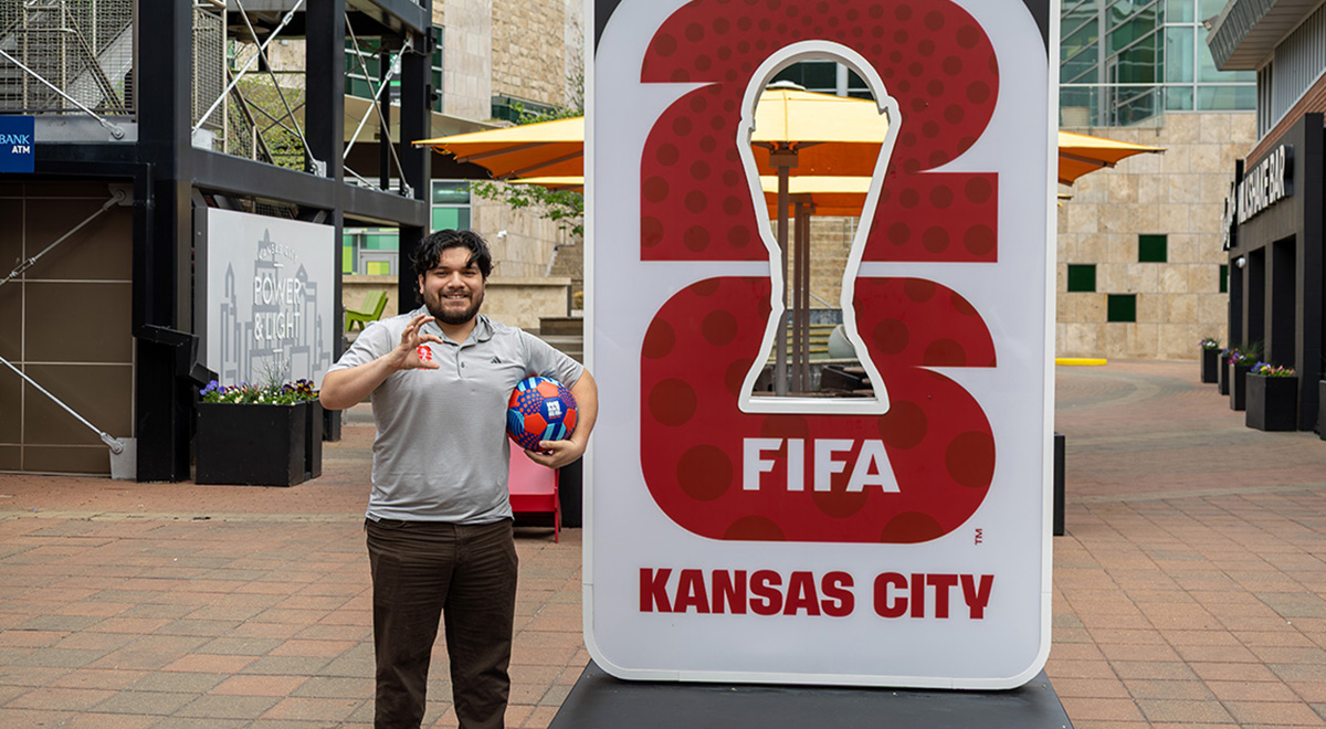A man with dark hair and a beard holds a decorative soccer ball next to a FIFA Kansas City sign in an outdoor plaza.