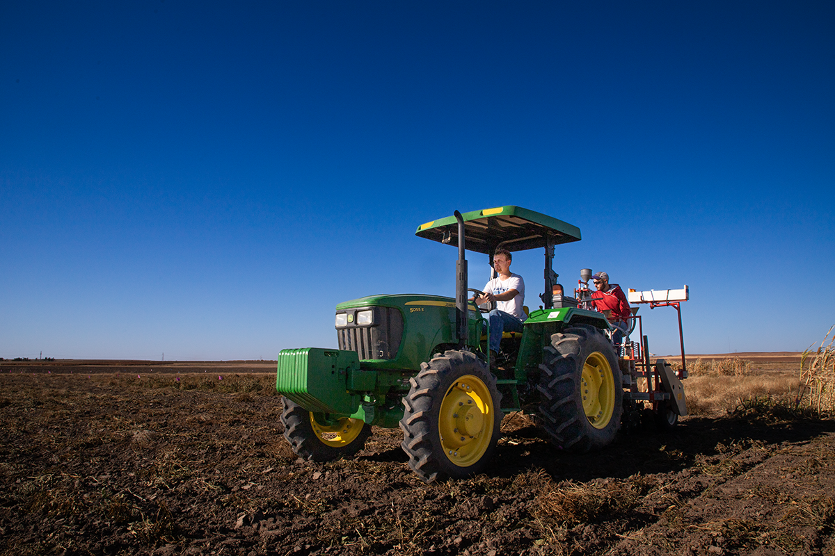 Two men ride a tractor in a brown field with a blue sky in the background