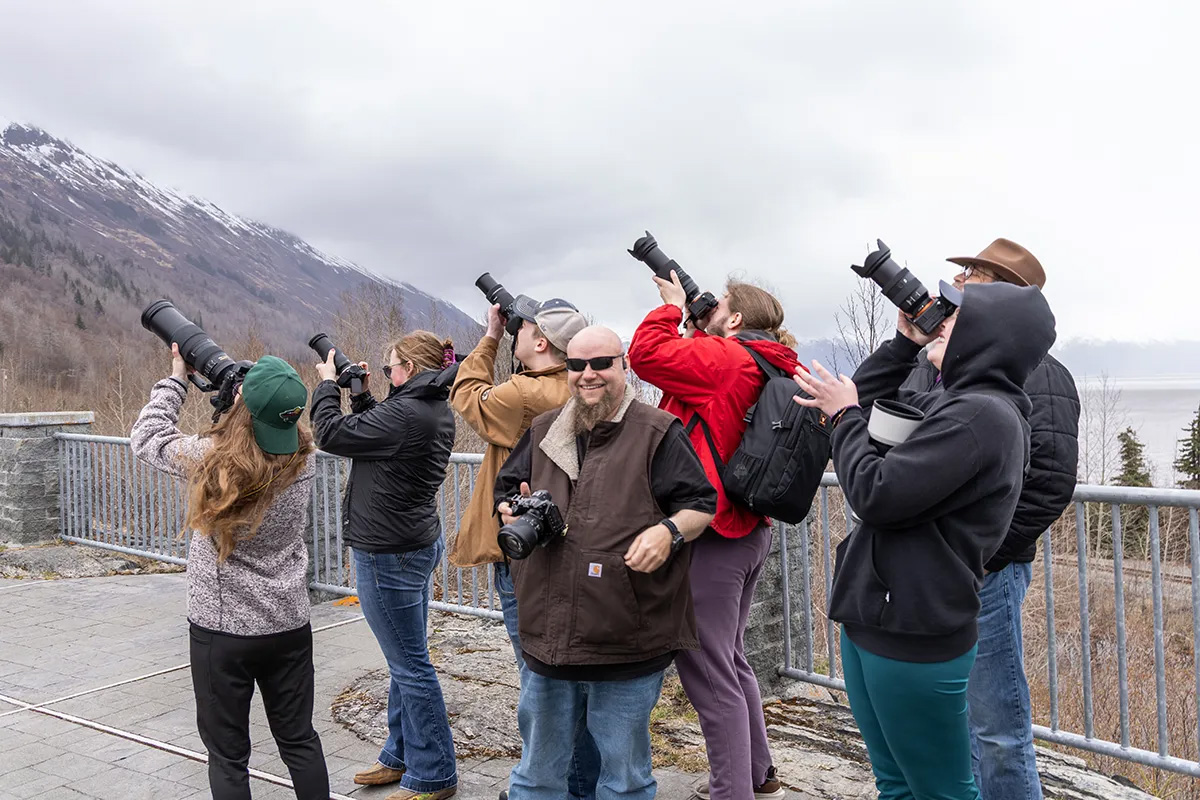 A group of college students all hold large cameras and aim them towards a focal point in the wilderness of Alaska. 