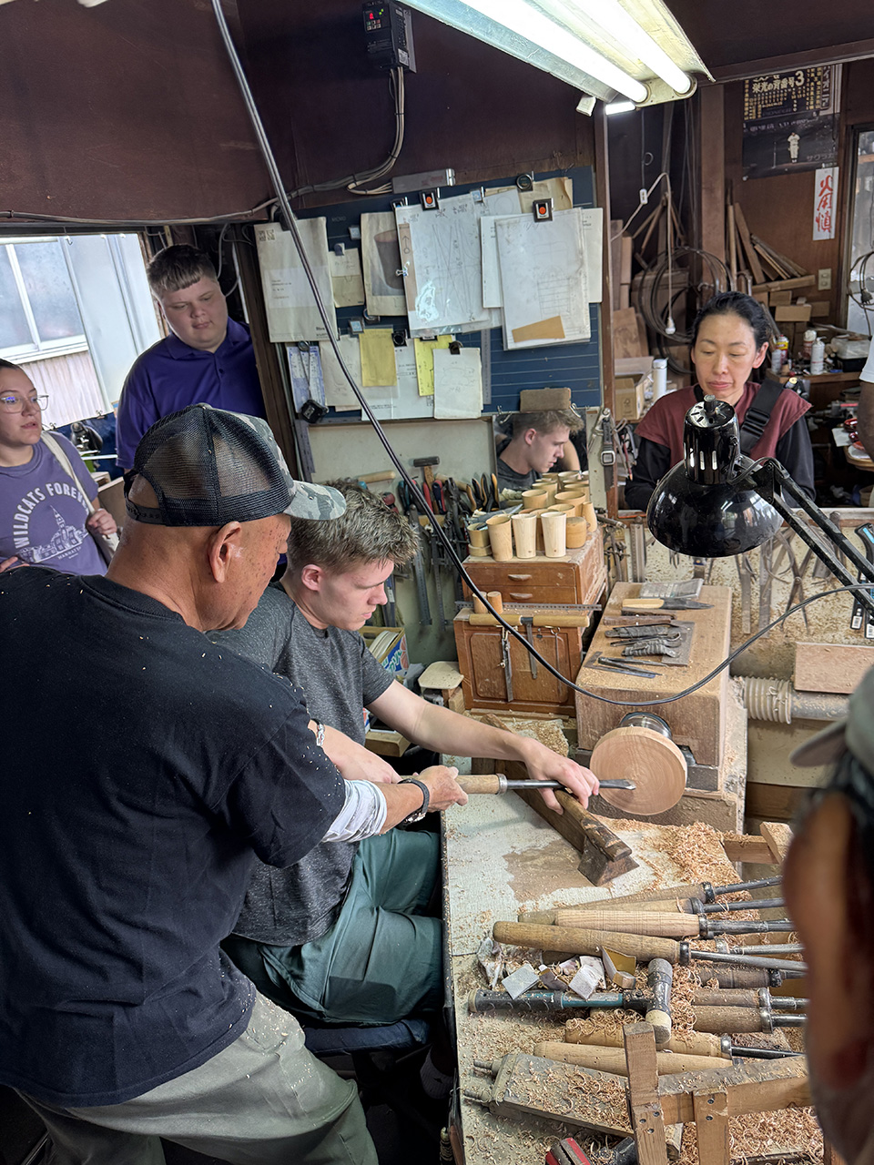 Students observe an artisan workshop. A man and a young man are holding a tool up to a circle of wood.