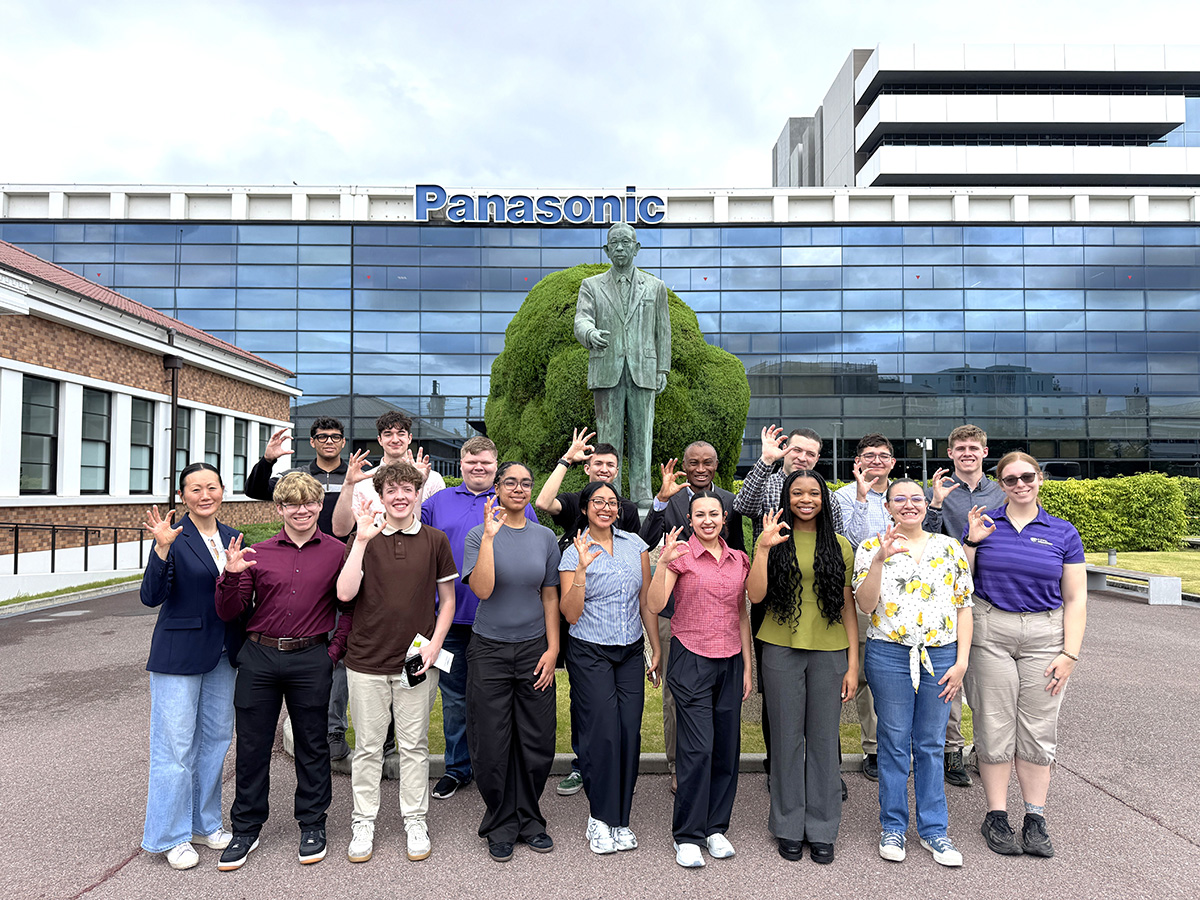 A group of people stand in front of a Panasonic building near a statue.