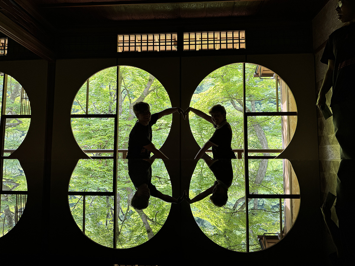 A dark photo is lit by two circular windows showing bright greenery outside, with the reflection on a table. Framed in two of the circles, two students use their arms to make a heart.
