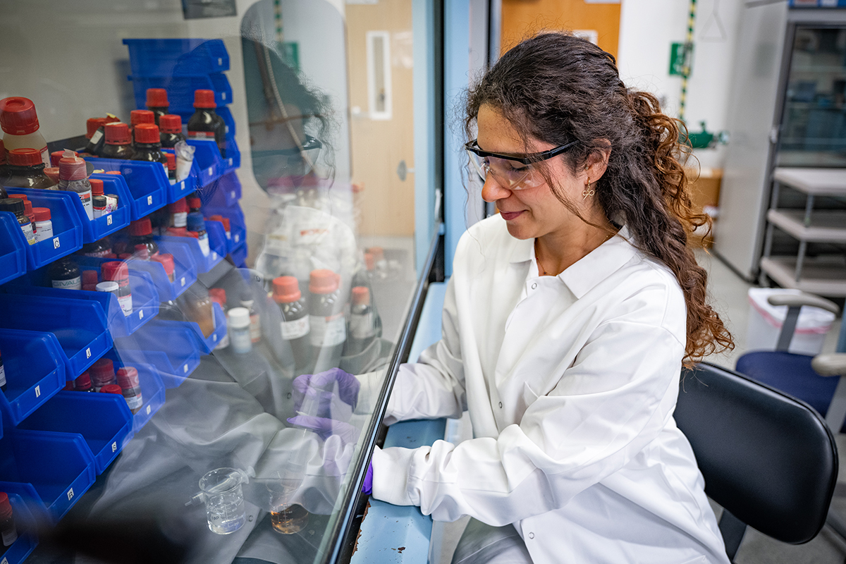 A female graduate student in a labcoat and safety glasses works with jars of liquid behind a research hood.