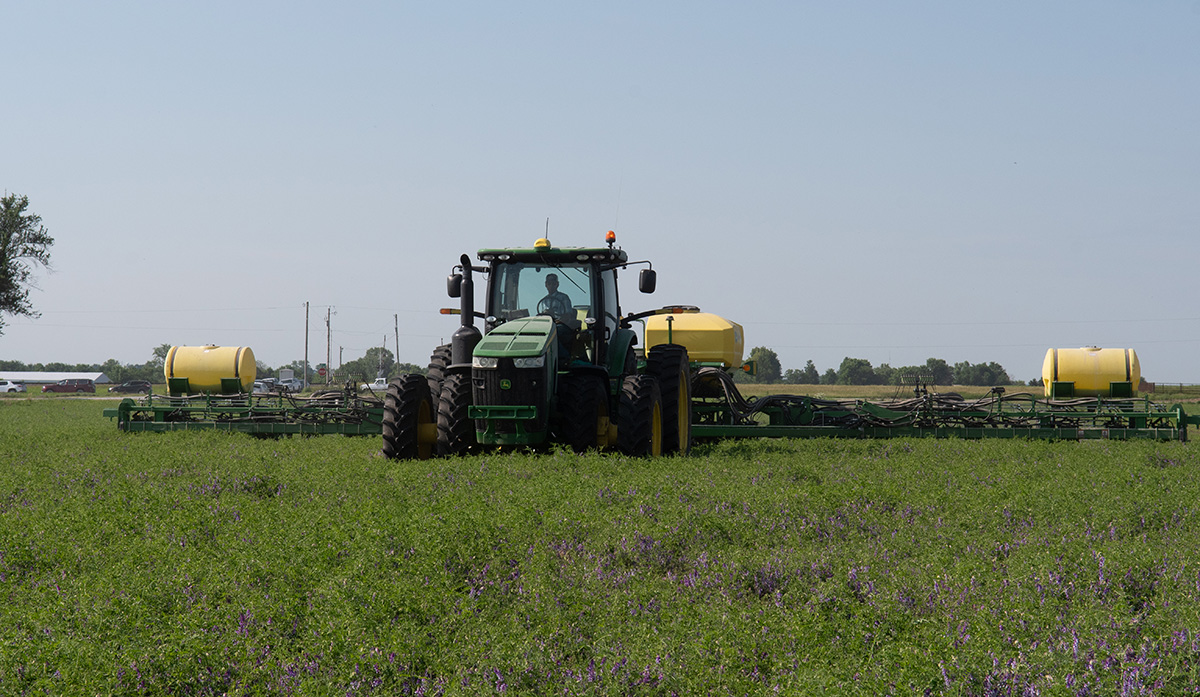 A tractor is pulling equipment on a green field with a few purple flowers 