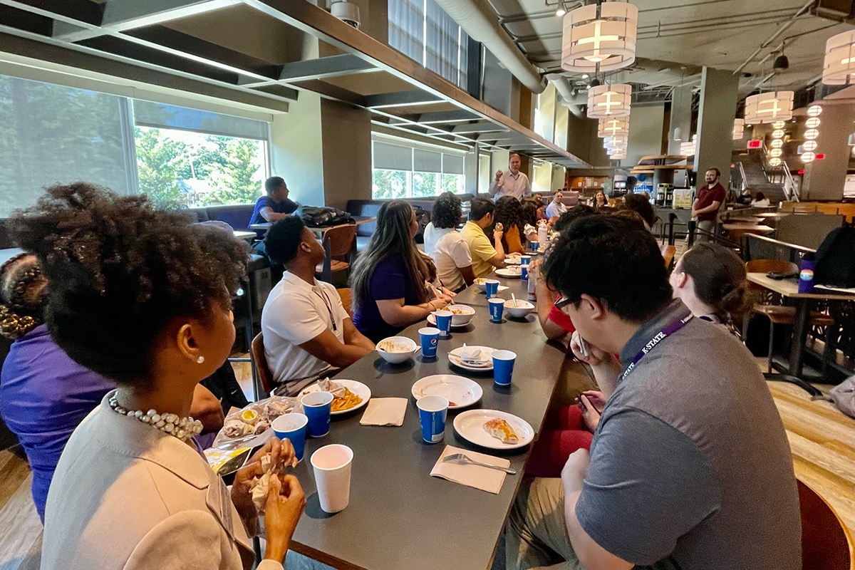 A group of high school campers sit at a long table in a college dining center.