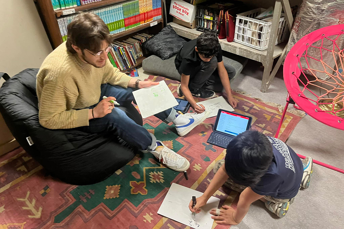 A male college student sits in a beanbag chair next to two elementary school-age boys who are sitting on the floor coloring.
