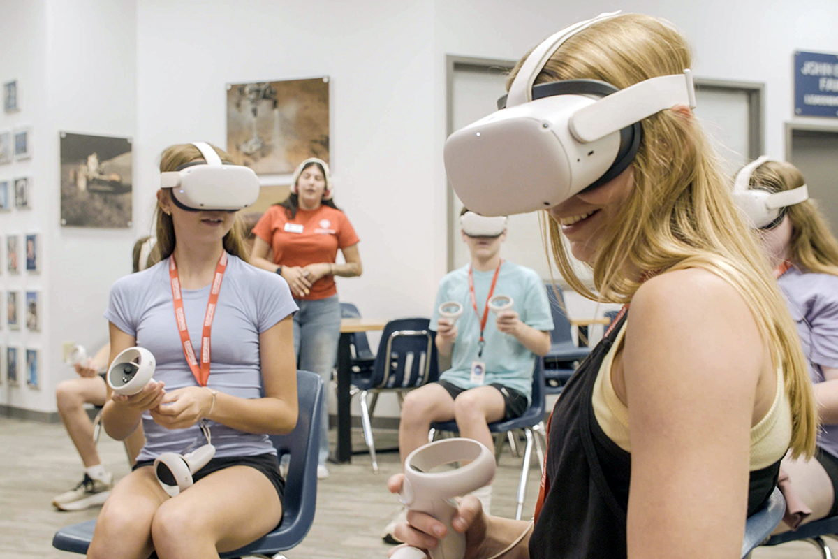 A couple of high school students wear and try virtual reality goggles at a space museum.