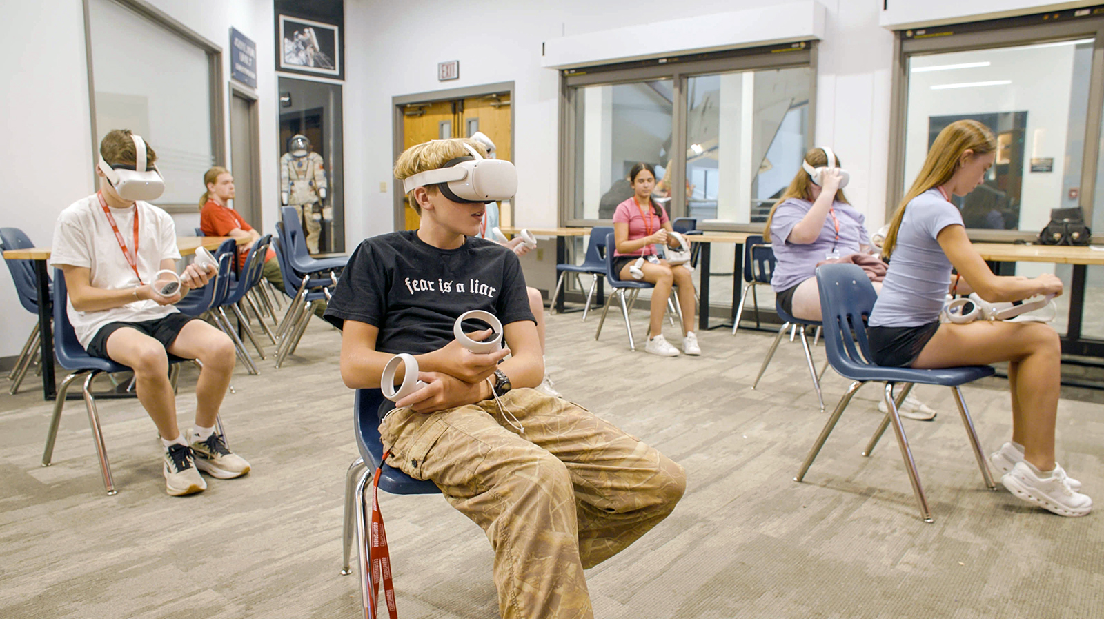 A group of high school students sit in a large classroom. Each is wearing a VR headset and interacting with a virtual reality game.