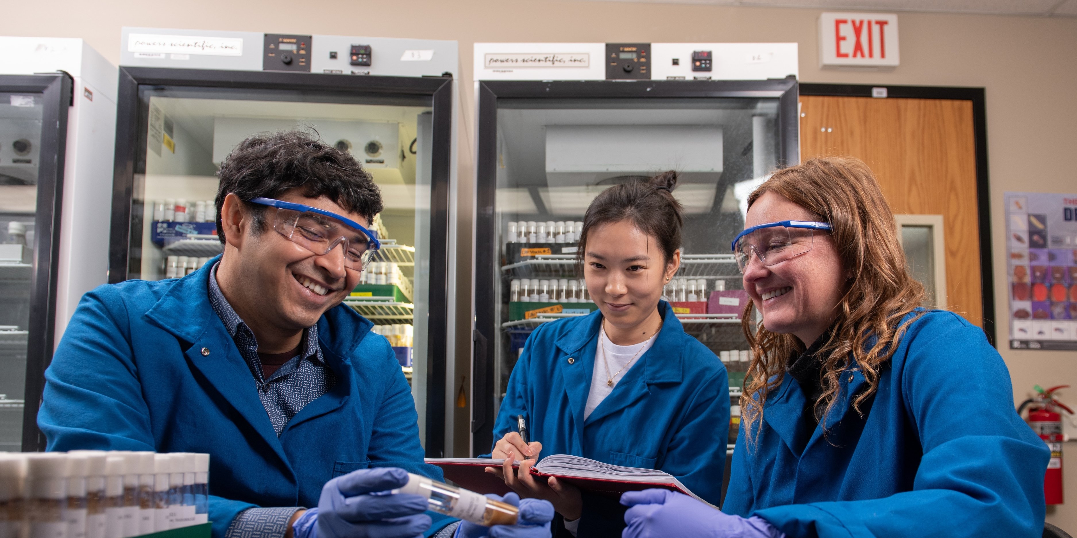 Geisbrecht analyzes lab samples with two students. All three wear blue lab coats and goggles.