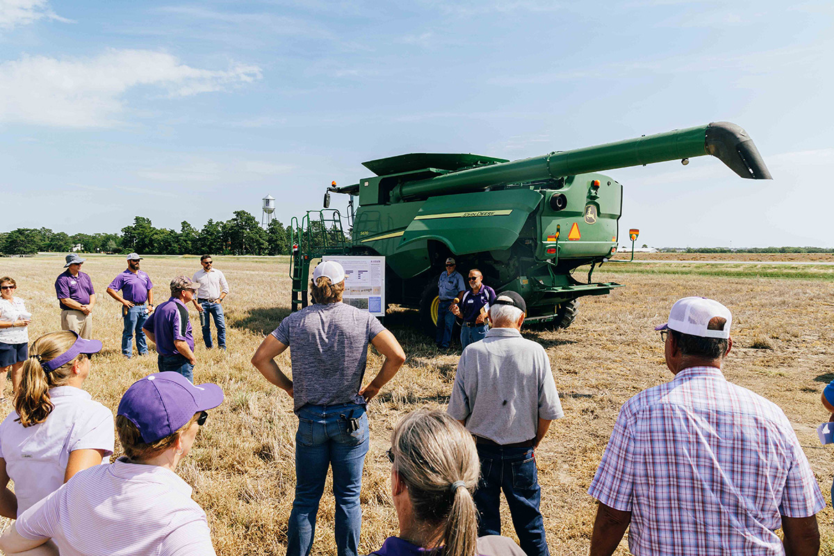 About 15 people stand in the middle of a field near a green combine.
