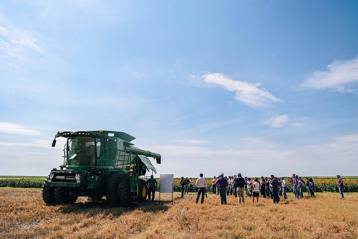 A green combine sits in a field with a group of people observing on one side under a big blue sky.