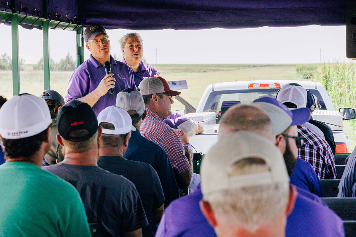 A photo from the back of a tour bus shows the backs of participants and two people in purple polos at the front speaking.