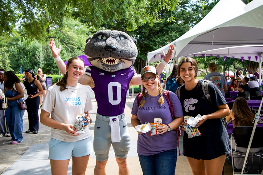 Willie the Wildcat stands with arms raised next to three students at an outdoor event