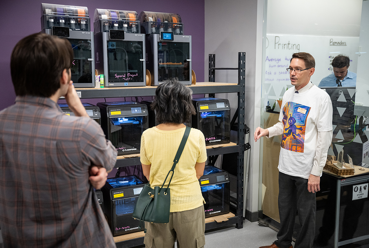 A man in a white long-sleeved shirt speaks to a woman in a yellow shirt in front of some 3D printers