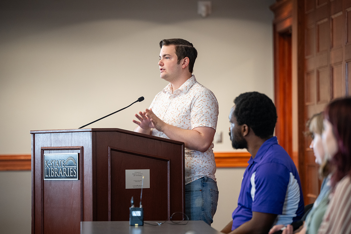 A student wearing a white shirt stands at a podium to present with other students looking on.