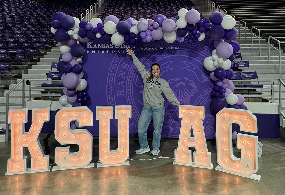 A students wearing jeans and a grey sweater stands in front of a purple background with a balloon arch between big light-up letters that say KSU AG.