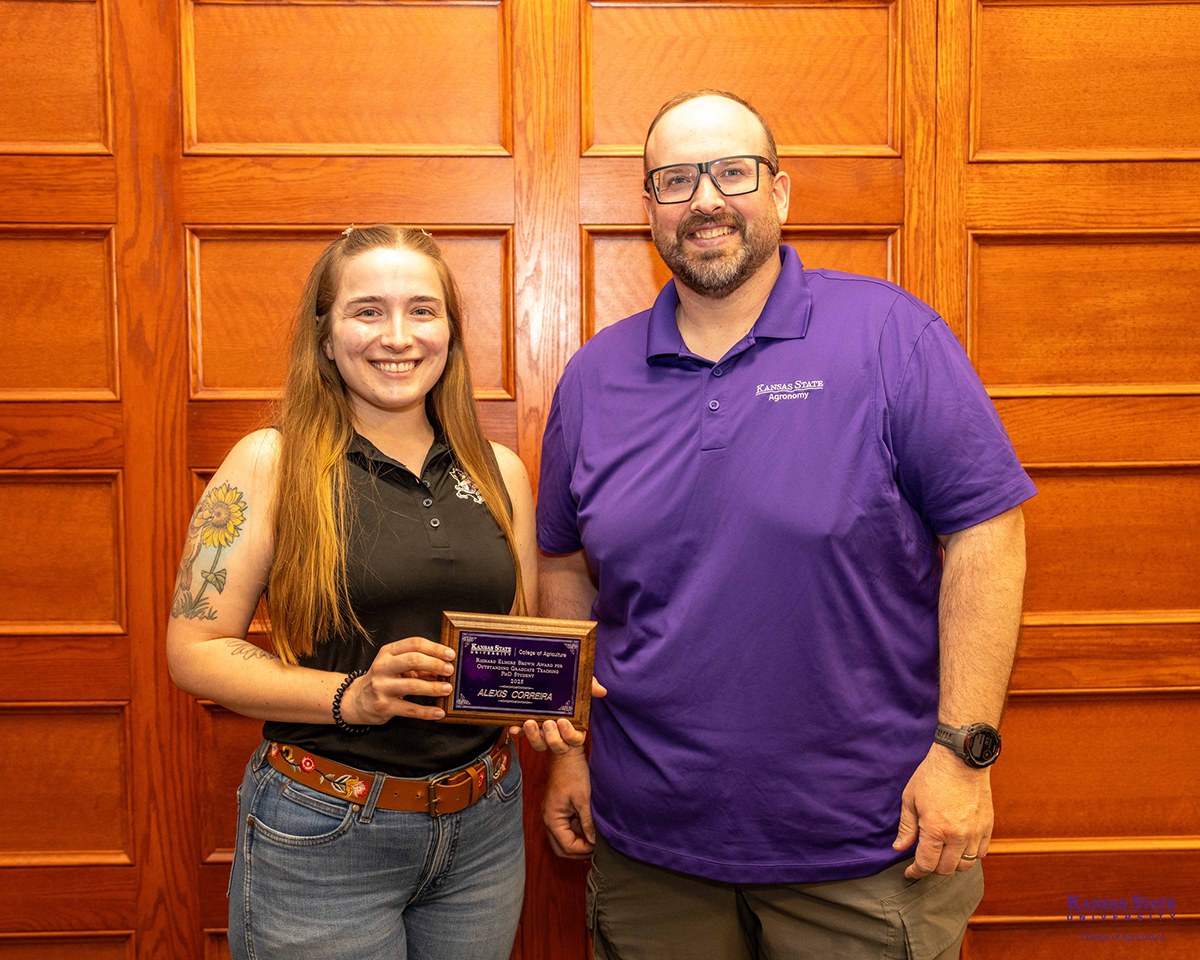 A student wearing a black top and blue jeans holds an award and smiles next to a man wearing a purple K-State agronomy shirt.