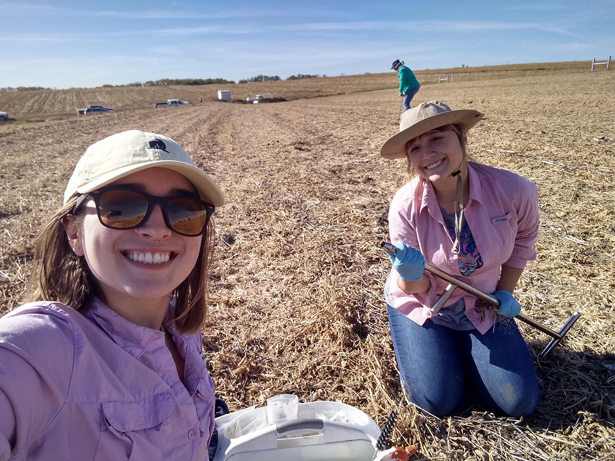 Two students wearing hats sit in a field holding rakes.