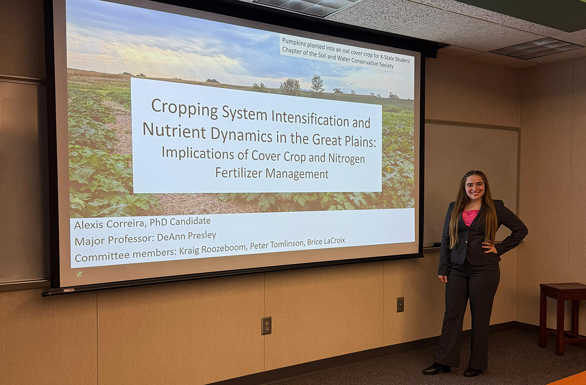 A student in a suit stands in front of a presentation screen with the title Cropping System Intensification and Nutrient Dynamics in the Great Plains: Implications of Cover Crop and Nitrogen Fertilizer Management.