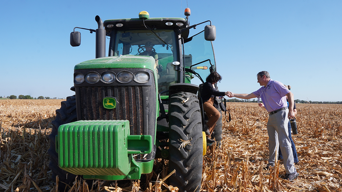 A man in a lavender polo helps a woman out of a tractor in the middle of a field.