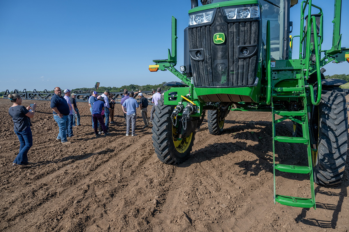 A large green piece of farm equipment sits in a dirt field with people gathered to its left.