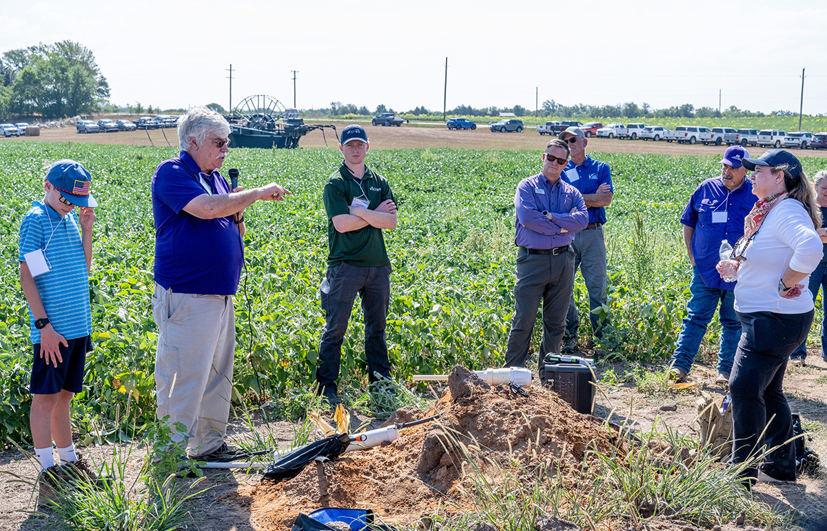 A man in a purple polo holds a microphone and speaks to a group of people on the outskirts of a field with green growth.