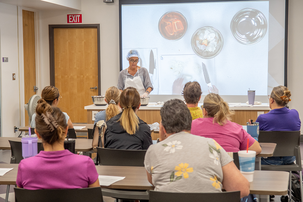 A food science professor in a hairnet presents to a class of Extension agents inside a kitchen classroom.