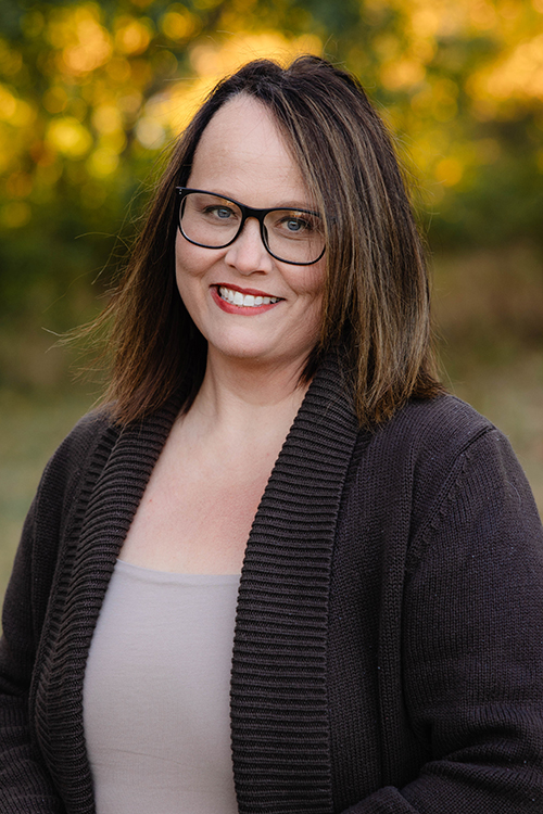 A woman in a black cardigan is pictured outside with green and yellow foliage blurred in the background.