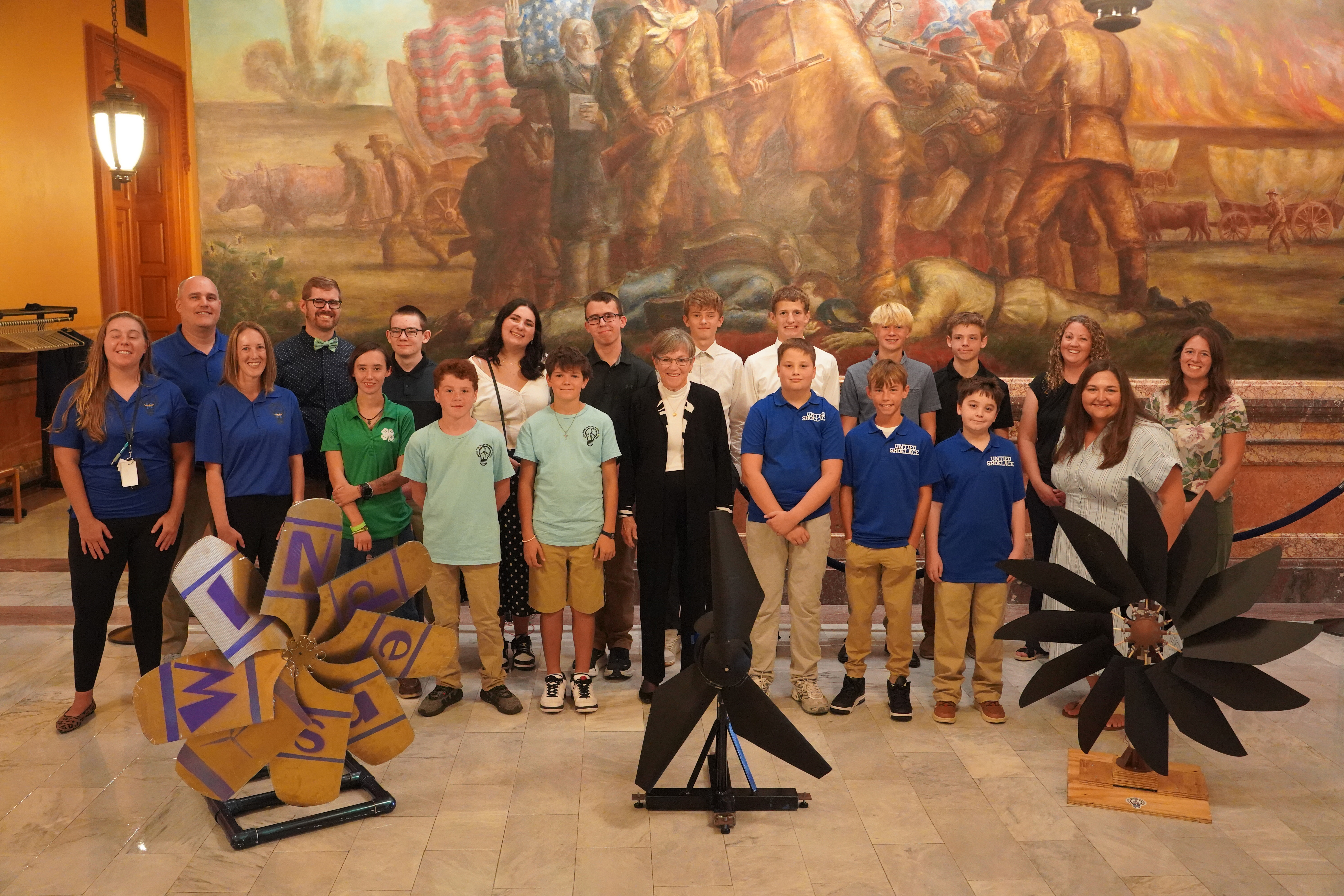 : A group of K-12 students stands with Kansas Governor Laura Kelly in the Kansas State Capitol alongside their winning wind turbine designs.
