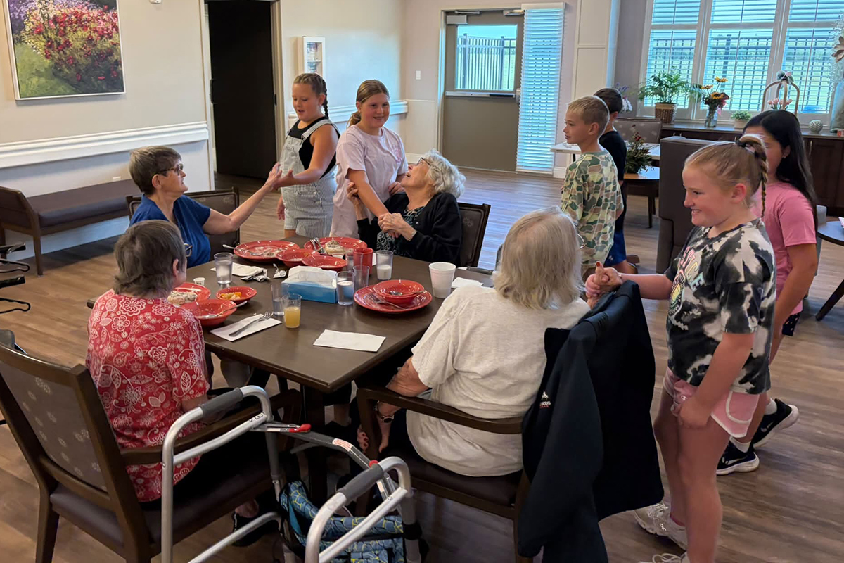 A group of young elementary school children interact with senior citizens at a breakfast table in a nursing home.