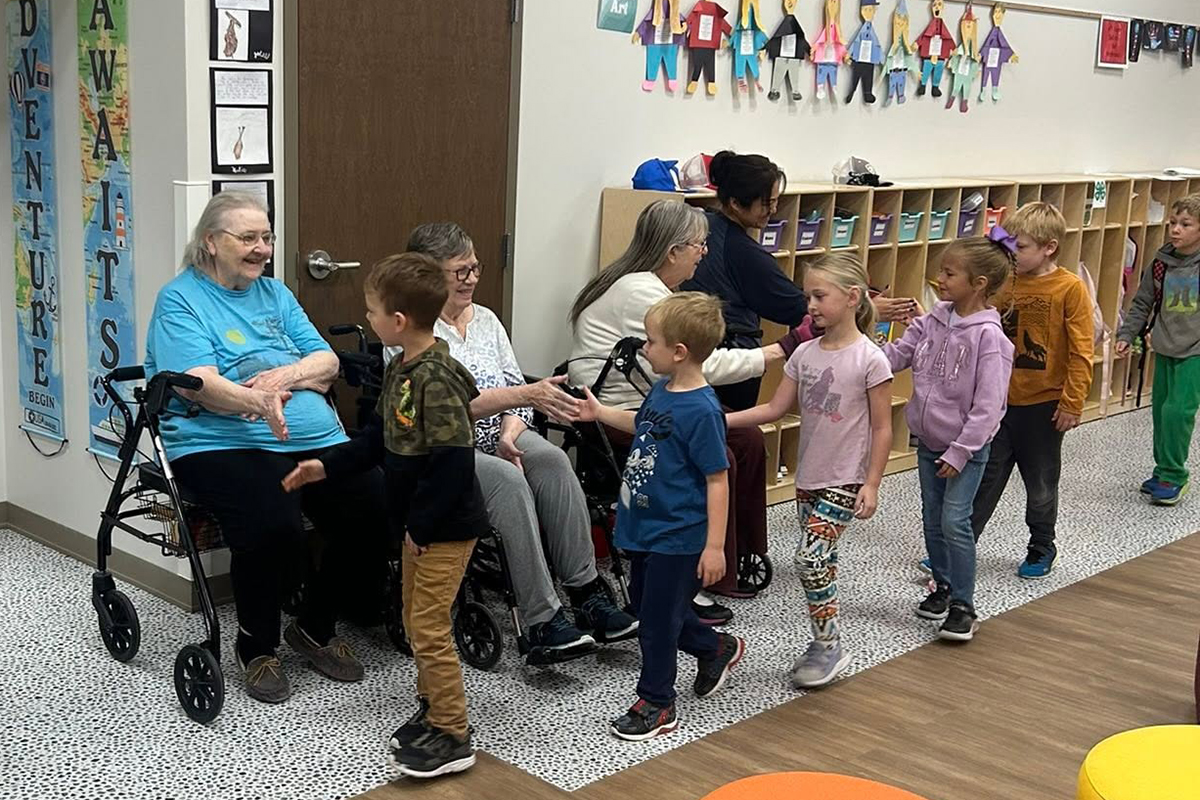 A group of young elementary school students walk down a hallway and high-five senior citizens of a nursing home.