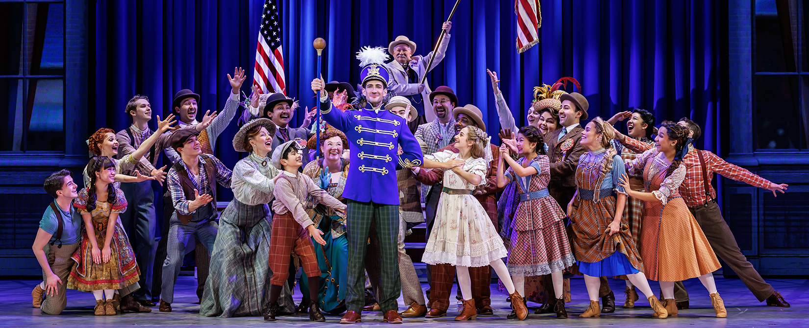 A stage production of The Music Man is pictured with the cast gathered behind a man in a drum major uniform with two American flags being held in front of a blue curtain.