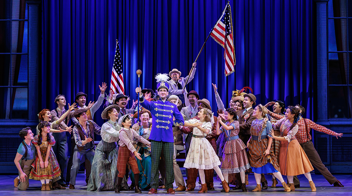 A stage production of The Music Man is pictured with the cast gathered behind a man in a drum major uniform with two American flags being held in front of a blue curtain.