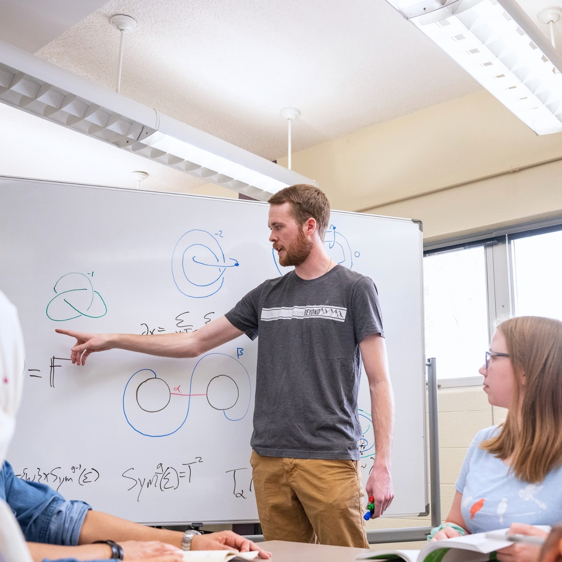 Jesse Osnes points at a whiteboard while explaining a mathematical solution to a table of college-aged students.