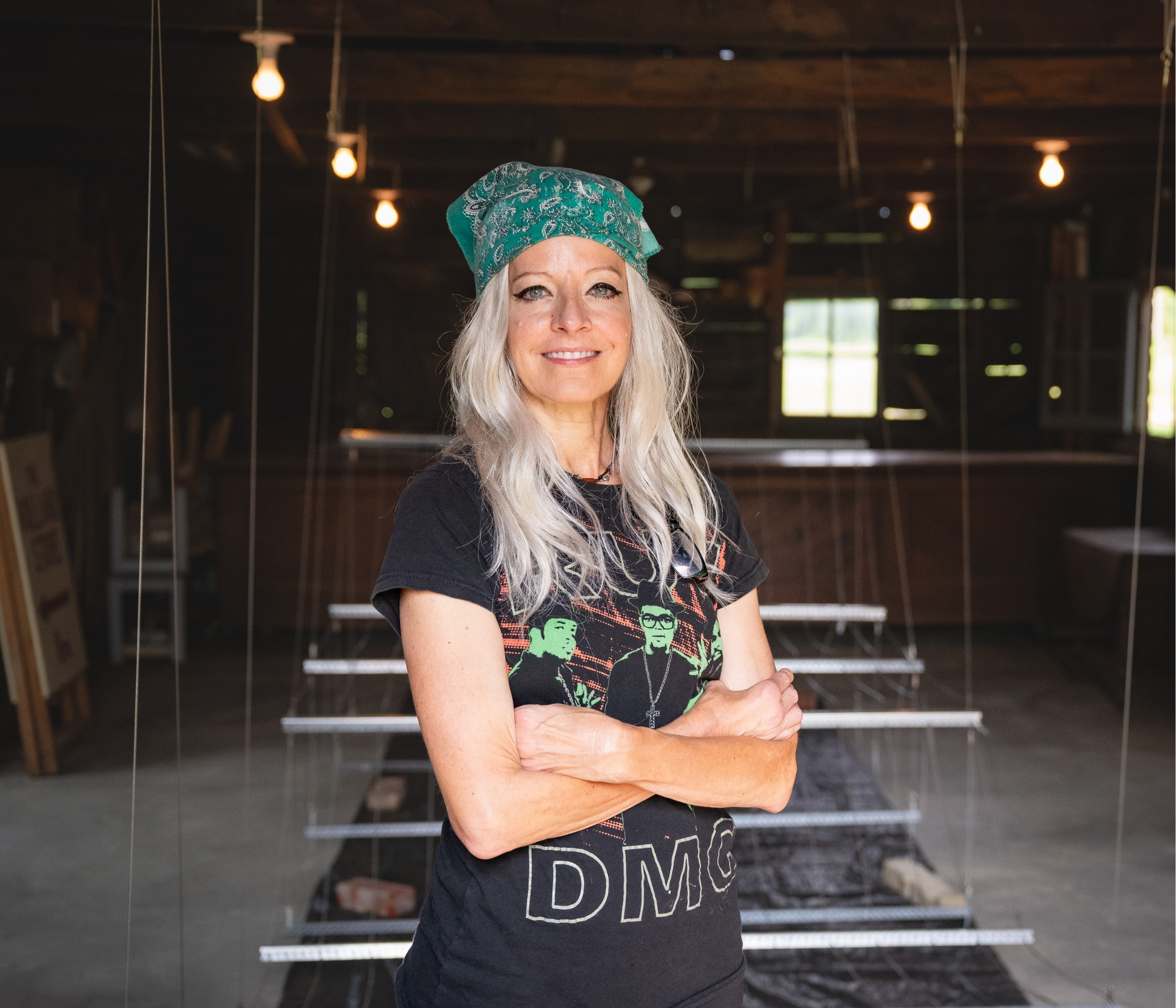 Ursula Emery-McClure stands in a barn, wearing a black t-shirt and green bandana. In the background is the hanging wire apparatus used for her project, 'The ROOT Source'