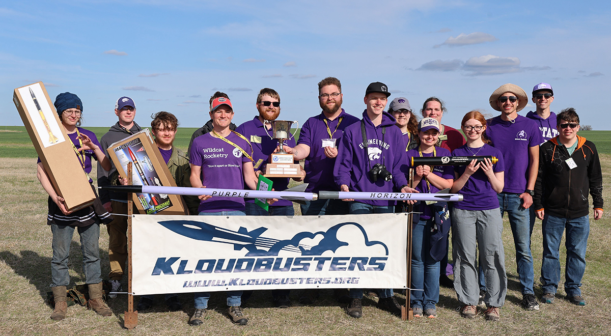 A group of students in purple shirts standing outside with rockets in front of a banner for Kloudbusters holding a trophy